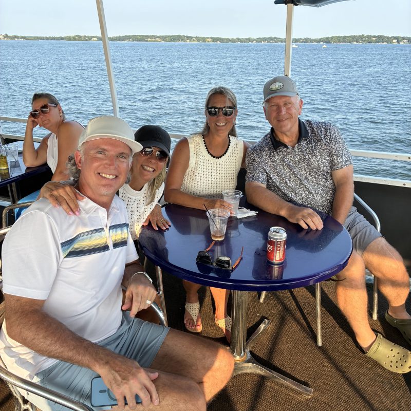 Five people sitting at a boat table with drinks, smiling, and a water view in the background.